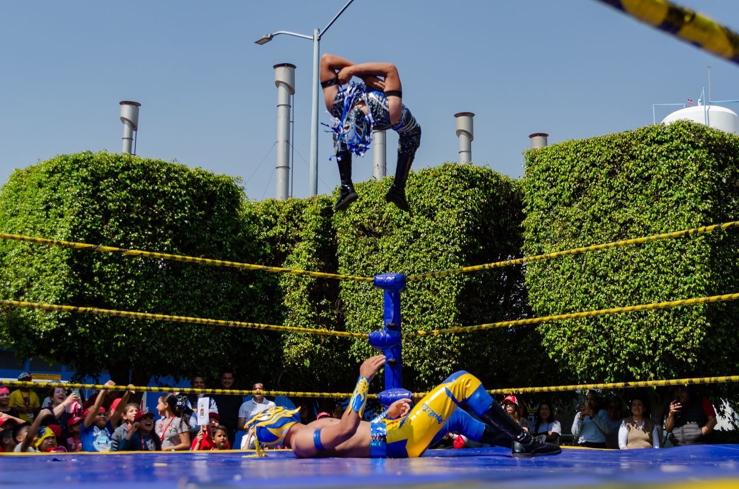 Luchador en el aire sobre el ring mientras otro espera en la lona y el público observa