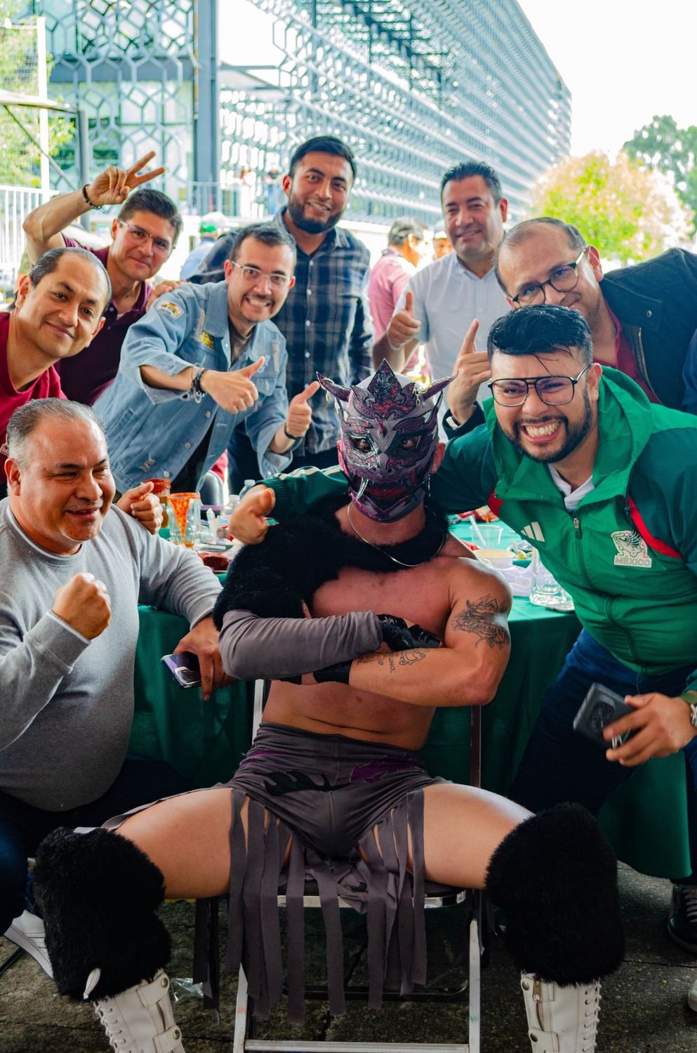 Grupo de asistentes sonriendo en una foto con un luchador enmascarado sentado