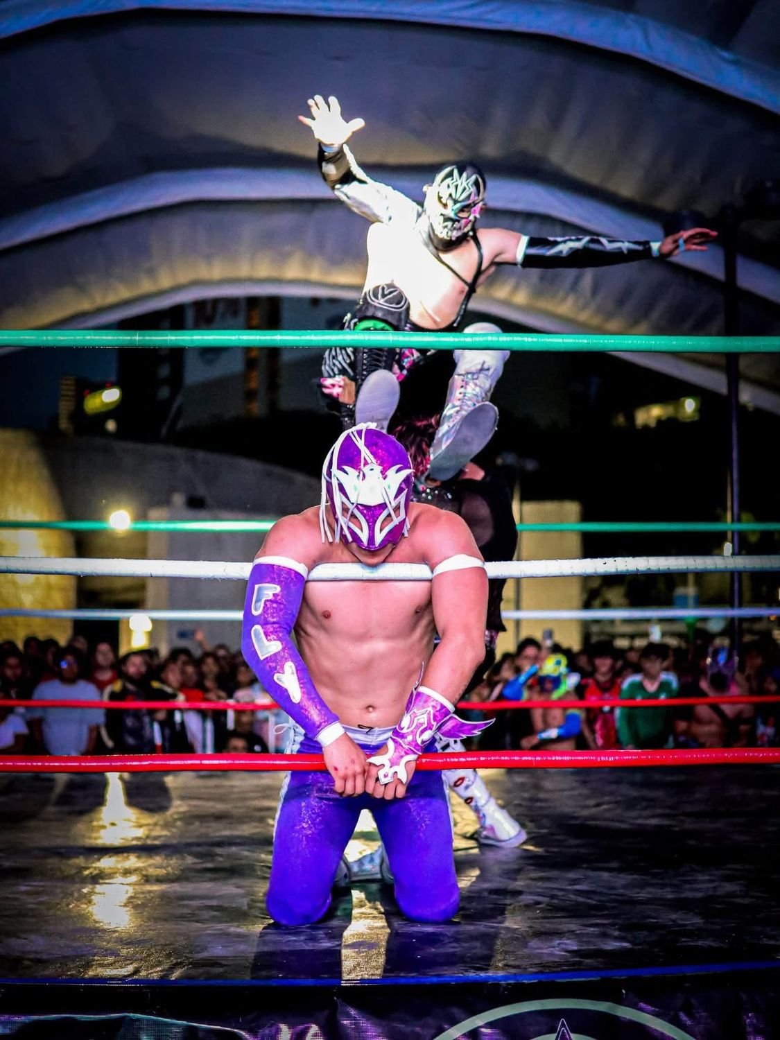 Luchador de máscara morada arrodillado en las cuerdas mientras otro salta detrás