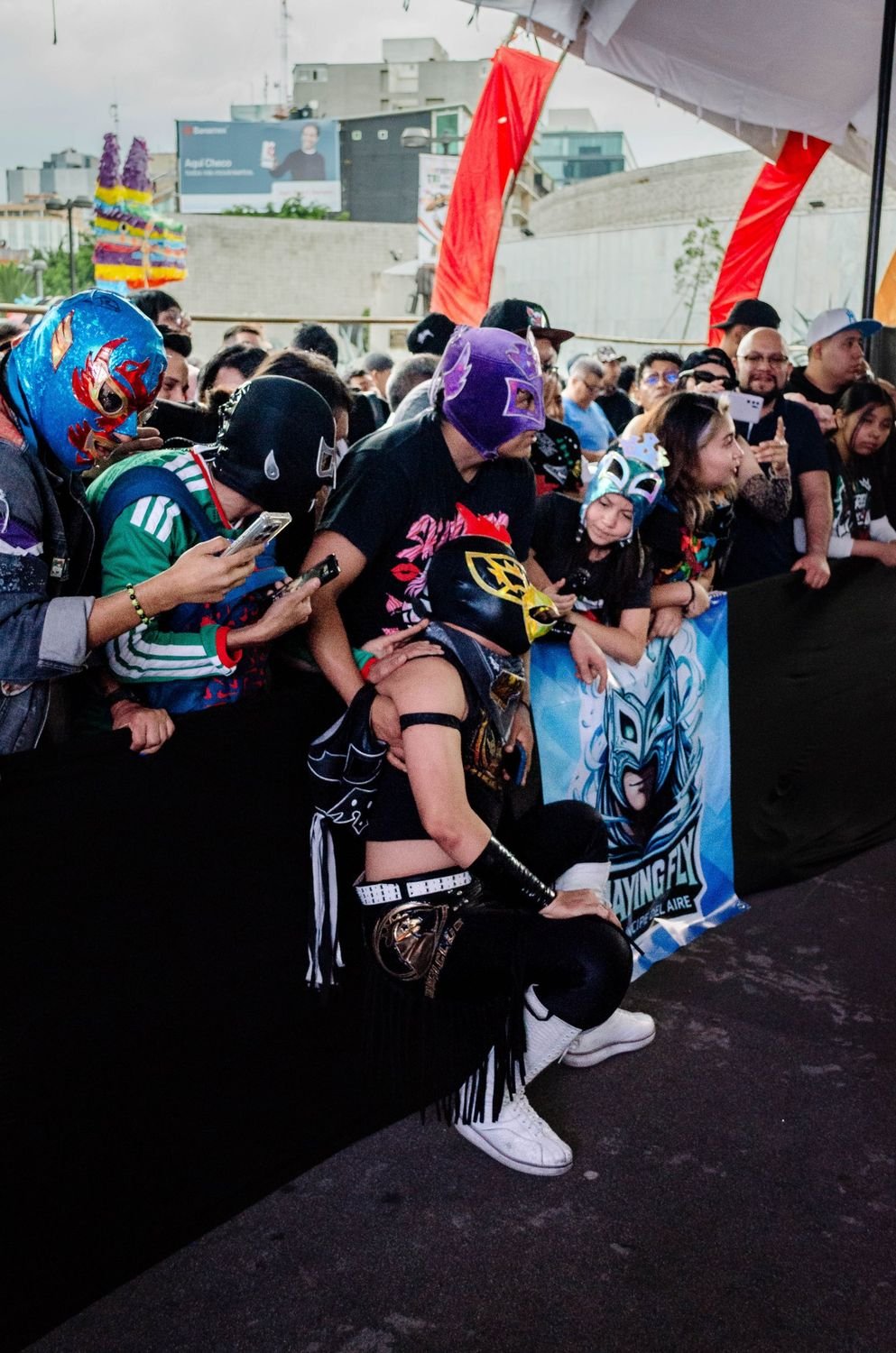 Aficionados con máscaras de lucha libre y celulares junto al ring mientras un luchador se agacha frente a ellos