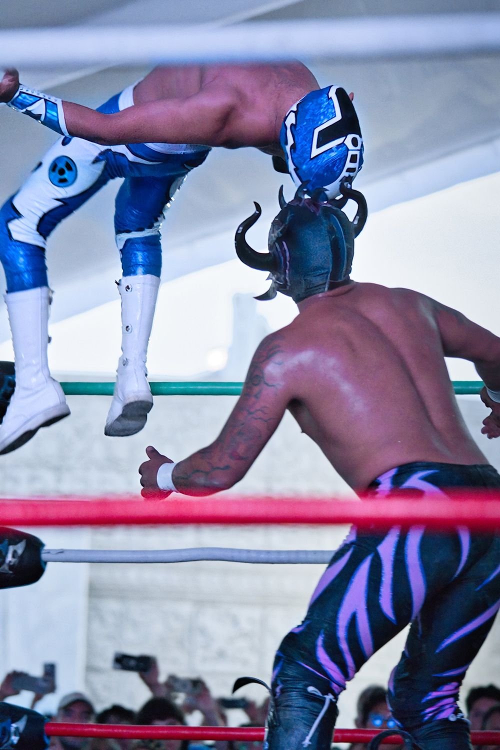 Luchador de máscara azul girando en el aire sobre un rival con máscara de cuernos