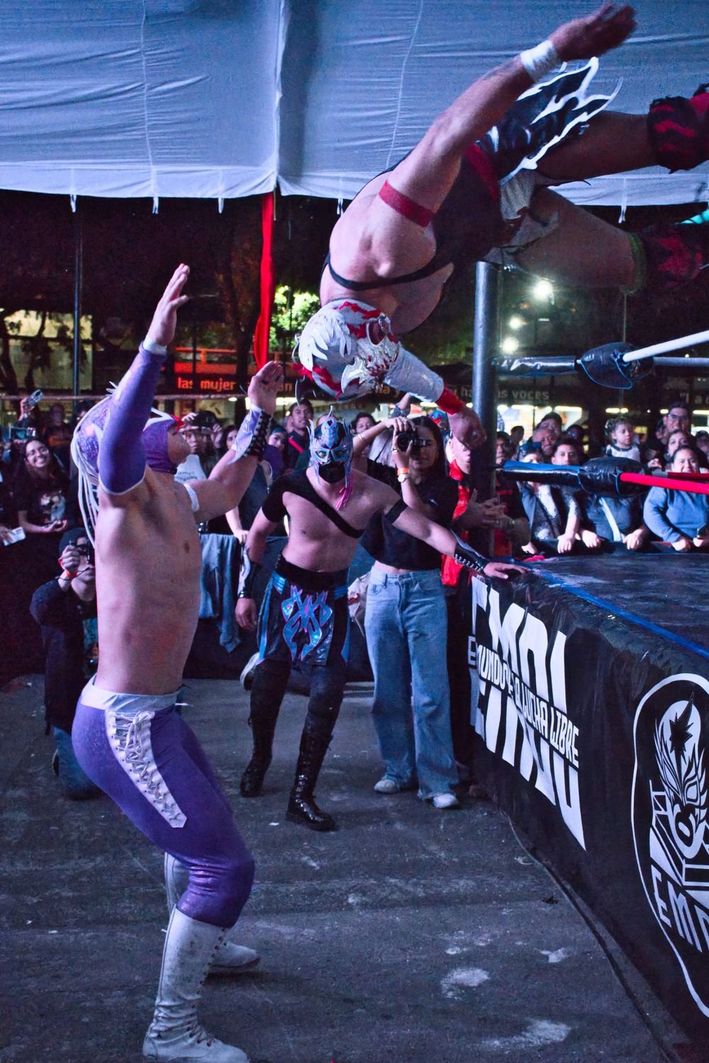 Luchador lanzándose desde las cuerdas junto a un ring con el faldón de El Mundo de la Lucha Libre, de noche